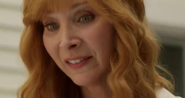 Lisa Kudrow stands outdoors in front of a white wooden house wall. She has her red hair long, down, style with a fringe and in waves. Lisa stars in a white blouse, matt foundation, dark pink matt lipstick and mascara.