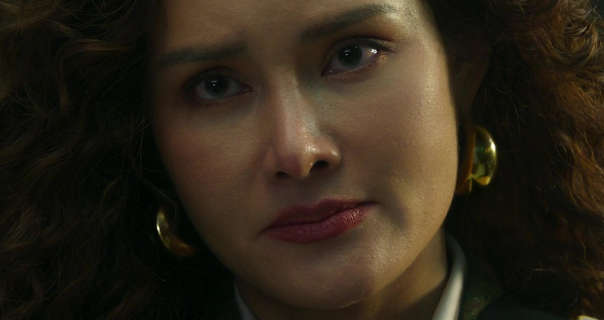 Rhatha Phongam stands with her long light brown curled hair filling the frame. She wears foundation with mascara, large gold earrings and a dark pink lipstick. She is lit from one side with a slightly pleading serious expression.