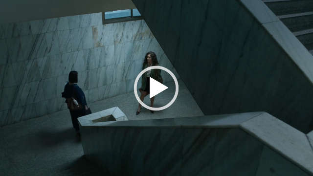 Rhatha Phongam stands and talks in a large white marble open stairway in flat daylight. She is seen in a grey blazer, skirt, black heels and a cream button up shirt. Rhatha has her long hair loose and in curls.