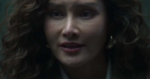 Rhatha Phongam is seen in a dark courtroom stairway. Her long hair is loose and styled in tight curls. Rhatha wears a light concealer with blush, pink lipstick and a pair of thick gold hoop earrings.
