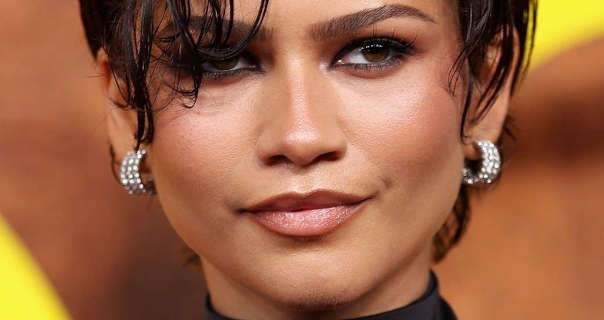 Zendaya is seen in front of a dark orange, brown and yellow poster wall at a television release event. She wears her short black hair styled messily with gel. She appears in a soft foundation, deep pink lipstick, black eyeliner and a pair if gemstone studded thick hoop earrings.