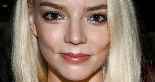 Anya Taylor-Joy is seen in a dark room at a television release event. Her long bright blonde hair is loose and straightened. Anya wears heavy mascara with a smoothing foundation and red lipstick.