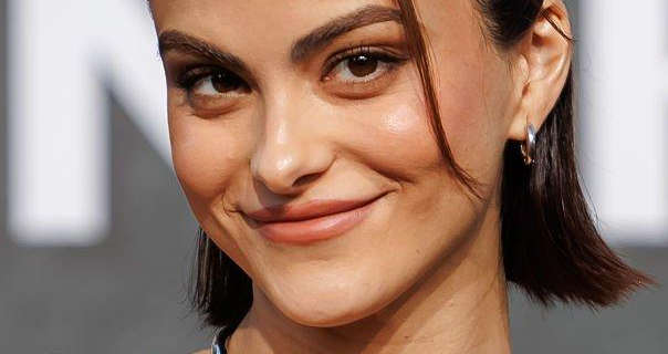 Camila Mendes smiles and looks into the camera. She stands in front of a light grey movie poster wall that has chunky white text across it. Camila has her black hair cut to just above shoulder length. She wears a matt foundation with blush, mascara pink lipstick and a pair of small silver hoop earrings.
