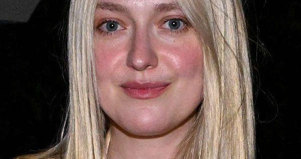 Dakota Fanning stands outside in the dark. Her face and straightened long blonde hair are light in a reflective bright light. Dakota wears a light foundation with blush, mascara and a dark pink sheen lipstick.