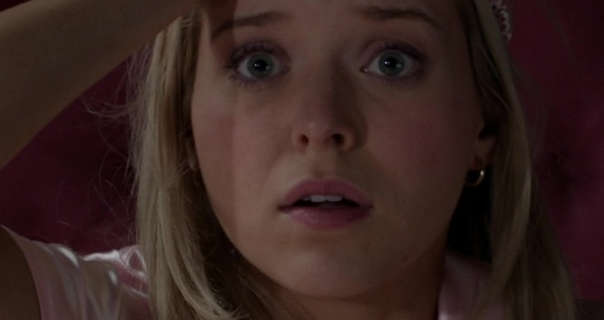 Lexi Minetree lifts and eye mask with one hand as she sits up in bed. She is seen with her blonde hair loose and straightened. Lexi wears a pink short sleeve pyjama top with matt foundation, blush, mascara, matt lipstick and a pair of small gold hoop earrings.