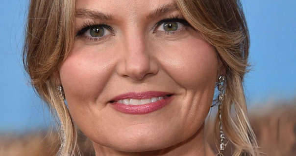 Jennifer Morrison stands in front of an event wall that has bright blue sky and marram grass on it. She wears her long blonde hair tie back in a tight bun with some strands left to frame her face. Jennifer is seen in a soft focus. She has a natural foundation with mascara, dark pink sheen lipstick and a pair of long hanging silver earrings.
