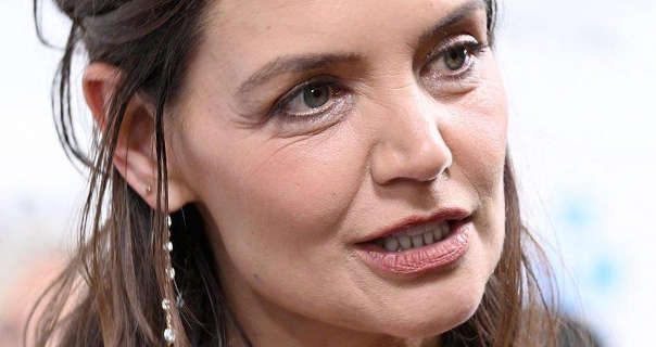Katie Holmes is interviewed in front of a white event wall. Katie has her long brown hair loose and pinned back. She wears a smoothing foundation with mascara, dark pink lipstick and a pair of hanging gemstone bead earrings.