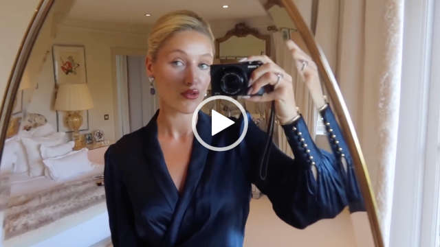 Leonora Smee films herself through a gold frame full length oval mirror in a bedroom at her countryside home. She wears a navy blue wrap dress with foundation, mascara, dark pink lipstick and a pair of large pendant earrings. Leonora has her long blonde hair tied back in a bun.