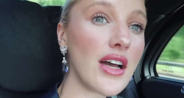 Leonora Smee leans forwards a little to look at her surroundings as she rides in the back of a Mercedes-AMG car to an event. Leonora has her long blonde hair tied back in a bun. She wears a matt foundation with pink blush, mascara, gloss lipstick and a pair of hanging glass bead with gemstone earrings.
