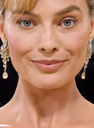 Margot Robbie looks straight ahead with a closed lip smile. She appears with her long hair tied back and up in a tight bun. Margot wears a smoothing foundation with mascara, dark pink lipstick and a pair of hanging gemstone earrings.