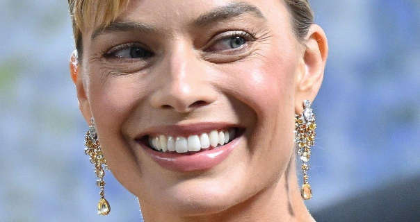Margot Robbie laughs and looks to the side. She is seen front of a blue, green and light black event wall. Margot has her blonde hair tied up and back in a bun. She wears a dewy foundation with mascara, pink gloss lipstick and a pair of hanging gold and silver chandelier gemstone earrings.