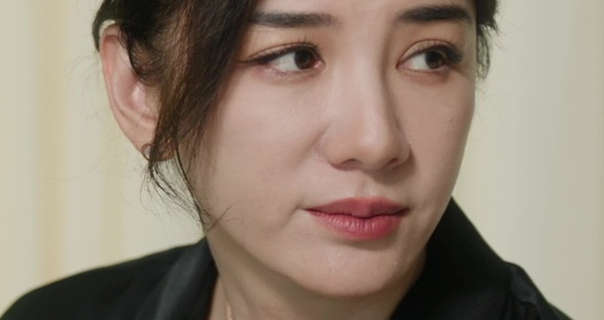 Huang Yi looks of to the side. She appears in front of a light closed hospital ward curtain. Huang Yi has her black hair tied up in a bun. She appears in a smoothing foundation, mascara, dark pink lipstick, gold jewellery and a black skirt.