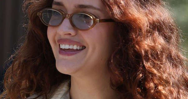 Olivia Cooke smiles and laughs as she walks outside in the London sunshine. She wears her long thick hair loose and in tight curls. Olivia appears in a pair of brown frame oval sunglasses. She wears a matt foundation with a dark red gloss lipstick and a light gold top.
