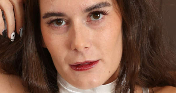 Louise B rests her head against her hand and looks into the camera. She wears a white halter neck dress with a light foundation, mascara, dark red lipstick and black and white painted nails. Her thick brown hair is loose and frames her face.