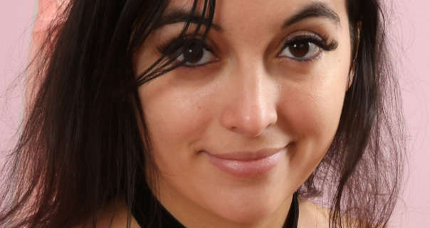 Summer Fox smiles a little as she looks into the camera. She is seen in front of a dusty pink wall. Summer has her thin black hair loose and in a natural style. She wears a light foundation with black eyeliner, heavy mascara and a dark pink sheen lipstick.