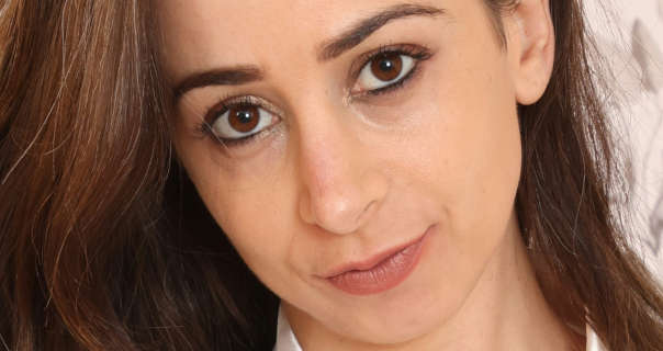 Tania Aresti looks into the camera while tilting her head slightly. She has her long and thick brown hair loose and pushed to one side. Tania is seen in a low coverage foundation, black eyeliner, mascara and a dark pink lipstick.