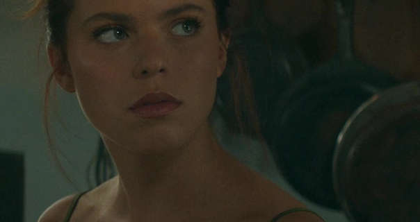 Heidi Grace Engerman is seen in an open plan kitchen at dawn. She glances upwards and to one side with a questioning look. Heidi has her long hair tied up in a messy bun. She wears a light coverage foundation with blush, curled lashes and a dark red matt lipstick.