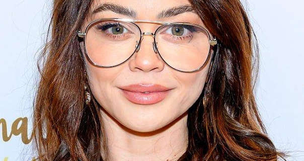 Sarah Hyland appears in front of a white event wall that has dark gold handwritten style text on it. Sarah has her brown hair loose and in messy waves. She wears a pair of large metal frame sunglasses with matt foundation, mascara, feathered eyebrows and a light pink lipstick.