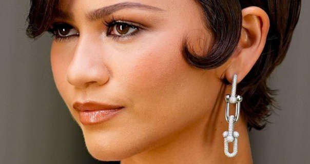 Zendaya is seen with her head turned looking into the distance. She has her black hair cut short and styled with a light gel. Zendaya wears a smooth foundation with blush, mascara, black eyeliner and a dark pink gloss lipstick. She stands outside in daylight and appears in a pair of large silver metal and diamond studded hanging chain earrings.