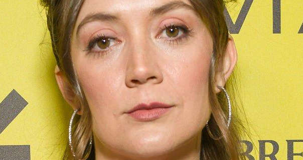 Billie Lourd looks straight into the camera as she stands in front of a yellow wall with dark grey logos and text across it. Billie has her long hair loose but pinned back. She wears a reflective foundation with blush, pink eye shadow, mascara, dark pink lipstick and a pair of large thin gemstone studded hoop earrings.