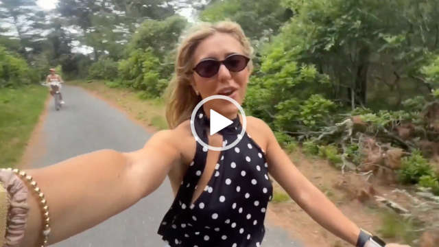 Em Sheldon cycles along a narrow tarmac path in an American woodland. Em wears a spotty black and white halter neck dress with her blonde hair loose and blowing in the wind. Em is seen in a pair of black frame sunglasses, foundation, pink lipstick, thin braclets and her Apple Watch.