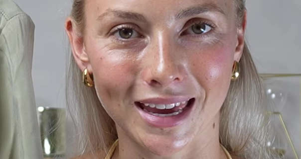 Emily Wilson Fashion sits in front of an off white wall with gold and glass ornaments on a shelf behind her. Emily has her long blonde hair loose and pinned back. She wears a pair of thick gold earrings with dewy foundation, pink blush, light mascara and a pink sheen lipstick.