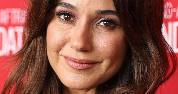 Emmanuelle Chriqui stands in front of a pastel red event wall that has white text across it. She has her long black hair loose, curled and with light highlights. Emmanuelle wears a natural foundation with mascara, black eyeliner and a dark pink satin lipstick.