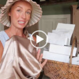 Josie Irons is seen in her stone kitchen at home near two white doors and a dark green wall. She appears in a pleated sun hat with her long hair tied in plaits. Josie wears a light foundation with pale pink lipstick and a thin necklace. She stands near a woven hamper full of new PR gifts. Josie holds up a gold hair care sleep covering.