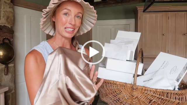 Josie Irons is seen in her stone kitchen at home near two white doors and a dark green wall. She appears in a pleated sun hat with her long hair tied in plaits. Josie wears a light foundation with pale pink lipstick and a thin necklace. She stands near a woven hamper full of new PR gifts. Josie holds up a gold hair care sleep covering.