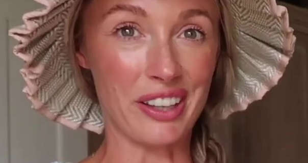 Josie Irons stands her kitchen at home in front of white cupboards and a dark beige wall. She wears a wide brim hat to help with the summer sunshine. Josie wears a light foundation, blush and a pink lipstick. Her hair is tied to the side in a single braid.