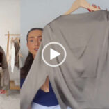 Hazel Maria Wood holds up a beige taupe colour long sleeve top on a light wooden hanger. She has her hair tied in a ponytail and is seen in a navy top as she stands in front of a dark white wall. Hazel wears a full foundation with mascara and a dark pink lipstick. There is a screen split to show her in the top and a pair of matching wide leg trousers.