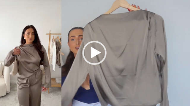 Hazel Maria Wood holds up a beige taupe colour long sleeve top on a light wooden hanger. She has her hair tied in a ponytail and is seen in a navy top as she stands in front of a dark white wall. Hazel wears a full foundation with mascara and a dark pink lipstick. There is a screen split to show her in the top and a pair of matching wide leg trousers.