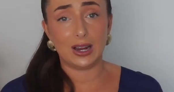 Hazel Maria Wood stands in front of a light grey wall. Hazel wears a shiny foundation with mascara, dark pink lipstick and a pair of large gold earrings. Hazel has her long hair tied back in a ponytail. She is seen in a thin navy long sleeve top.