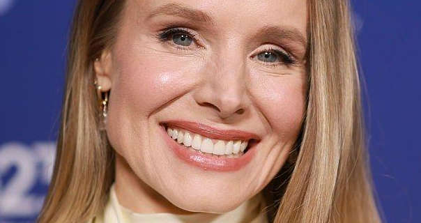 Kristen Bell stands in front of a purple wall with out of focus white text across it. She has her long blonde hair straightened and loose. Kristen wears a light coverage foundation with mascara, dark pink lipstick and a pair of dangling gold earrings.