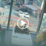 Kang Min-ah sits in front of a large glass office window with a busy street outside. She is seen from the side with her long black hair tied back in a bun. Kang Min-ah wears a black and red fluffy jacket with black knee high boots and soft makeup.