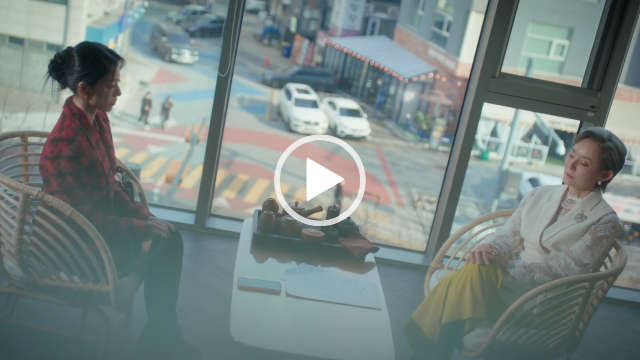 Kang Min-ah sits in front of a large glass office window with a busy street outside. She is seen from the side with her long black hair tied back in a bun. Kang Min-ah wears a black and red fluffy jacket with black knee high boots and soft makeup.
