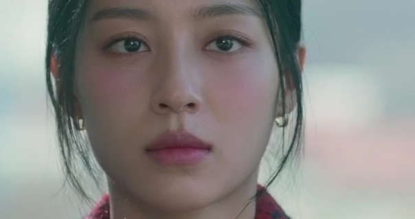Kang Min-ah looks forward with a blank stare. She has her long black hair tied up and back. Kang Min-ah wears blush over a matt foundation, pink lipstick and a pair of small gold hoop earrings.