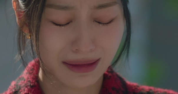 Kang Min-ah stands with her eyes closed and tears on her checks. She appears in a light office foyer. Kang Min-ah has her long black hair tied up. She wears a red and black fluffy blazer with a matt foundation, mascara and a pink lipstick.