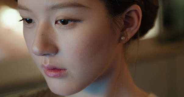 Zhao Lusi is seen close up as she sits in a study at night. She has her long black hair tied up in a bun. Lusi wears a smoothing foundation with black eyeliner, mascara, pale pink lipstick and a pair of pearl earrings.