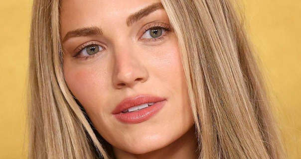 Michelle Randolph appears in front of a gold gradient wall. Michelle has her long blonde hair loose, straightened and curled inwards at the ends. She wears a full coverage foundation with mascara, soft blush and a pink lipstick.