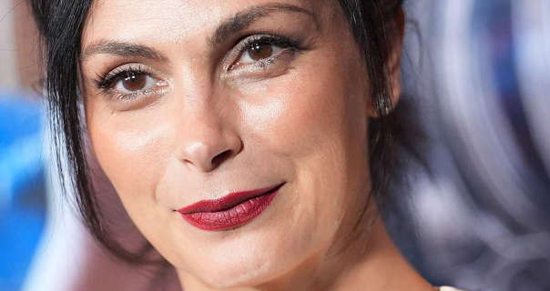 Morena Baccarin appears in front of a superhero movie poster wall at an event. She has her black hair tied back in a short ponytail. Morena wears a light coverage foundation with blush, curling mascara, deep red lipstick and a pair of gemstone earrings.