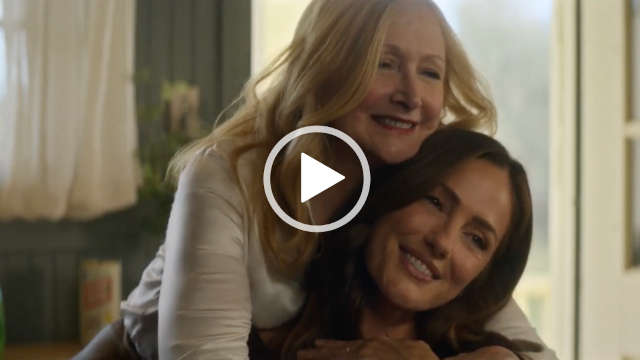 Patricia Clarkson leans down and hugs a smiling Minka Kelly. They appear in a rustic green and white wood kitchen near an open door. Patricia has her long hair loose and in soft waves. She wears a dewy foundation with red lipstick and black mascara.