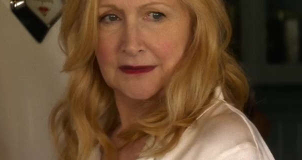Patricia Clarkson stands in an American country kitchen next to a large white classic fridge and dark green wood and glass cabinets. She wears her blonde orange hair loose and in soft curls. Patricia is seen in a light coverage foundation, pink blush, mascara and a deep red matt lipstick.