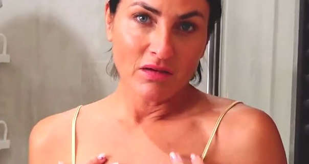 Sarah Wild looks into the camera as she stands in front of a chrome shower area. She has her black hair tied back. Sarah wears a light coverage foundation with mascara and a pink lipstick.