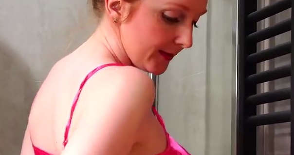 Miss Daphne Red appears in a cream tile bathroom near a metal heated towel radiator. She has her long hair tied up in a bun. Daphne wears pink strappy nightwear with small earrings, foundation, blush, mascara and a red lipstick.