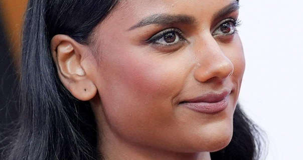 Simone Ashley is seen from one side as she stands in front of a white, dark orange and black event wall. She has her thick black hair loose and pushed back. Simone wears foundation with blush, mascara, black eyeliner, dark mauve lipstick and no earrings.