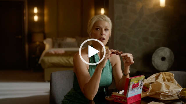 Hannah Waddingham sits on a grey chair at a large circular wooden table in an open plan hotel room. She has her blonde hair cut short and pushed back. Hannah wears a sleeveless green dress. She eats takeaway meat covered in a dark sauce.