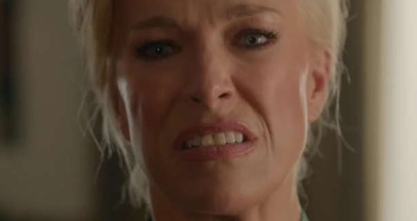 Hannah Waddingham scowls a little in disgust as she stands in a brown and beige hotel room. She has her long light blonde hair tied back. Hannah wears heavy mascara with a matt foundation and a dark pink lipstick.