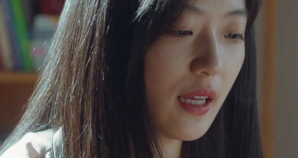 Sun Qian sits in front of a wooden bookcase with daylight hitting her back. She has her long, thick black hair loose and straight. Sun Qian wears foundation with black mascara, red lipstick and a pair of light pink pyjamas.