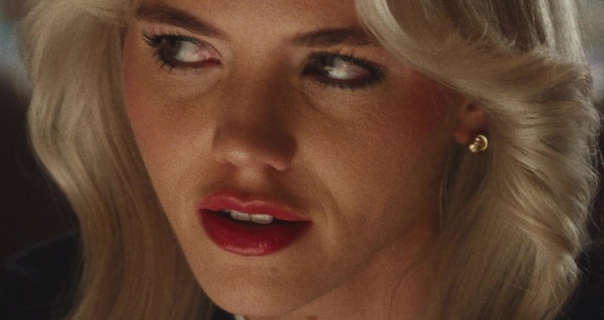 Hayes Warner glances up and to the side. She sits in a quite dark bar eating area. Hayes has her shoulder length blonde hair loose and layered. She wears a heavy foundation with blush, black clumping mascara, dark red gloss lipstick and a pair of small gold earrings.